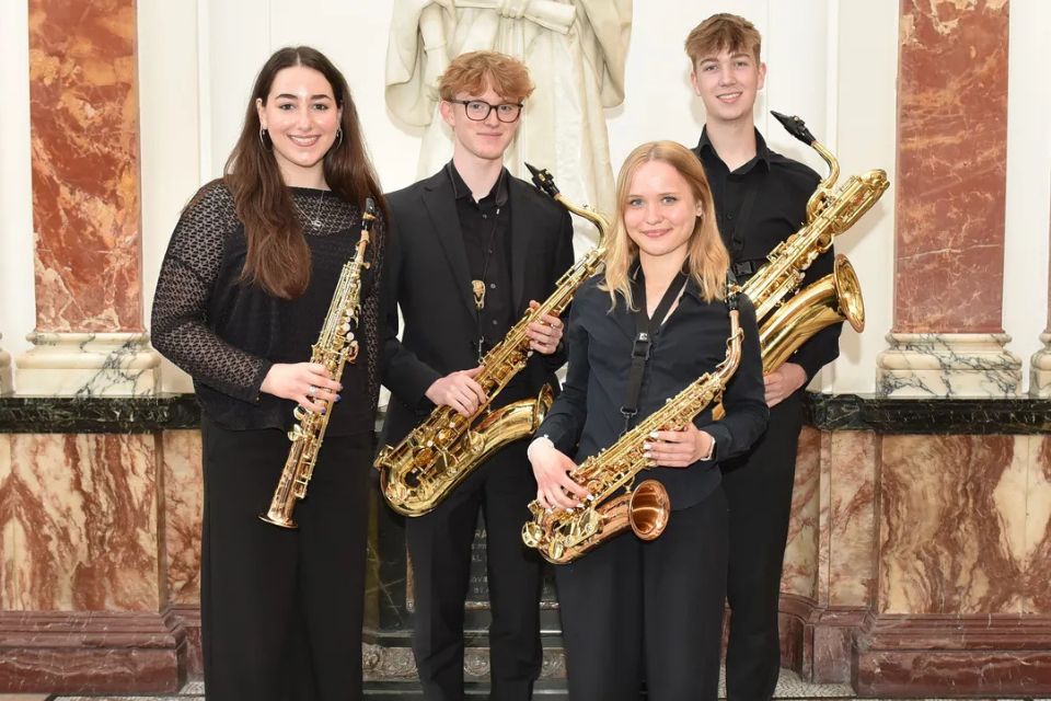 Kezia Colton and the RCMJD Saxophone Quartet wear black and hold their saxophones, smiling