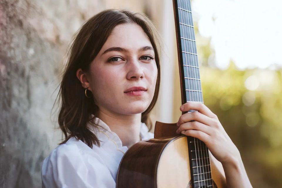 A young woman wears a white shirt and holds a guitar