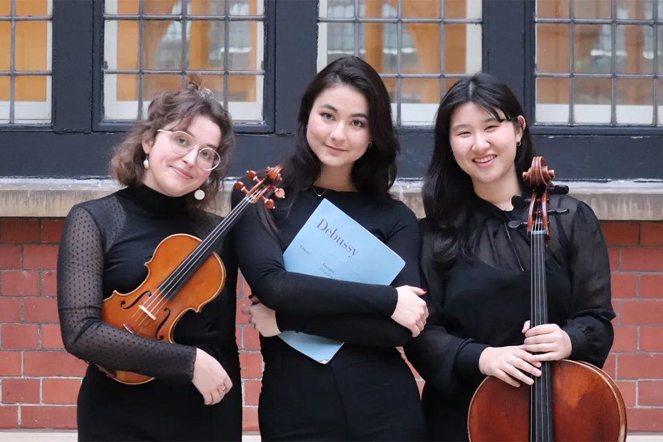 The Kaja Piano Trio wear black and hold their instruments in a courtyard, smiling