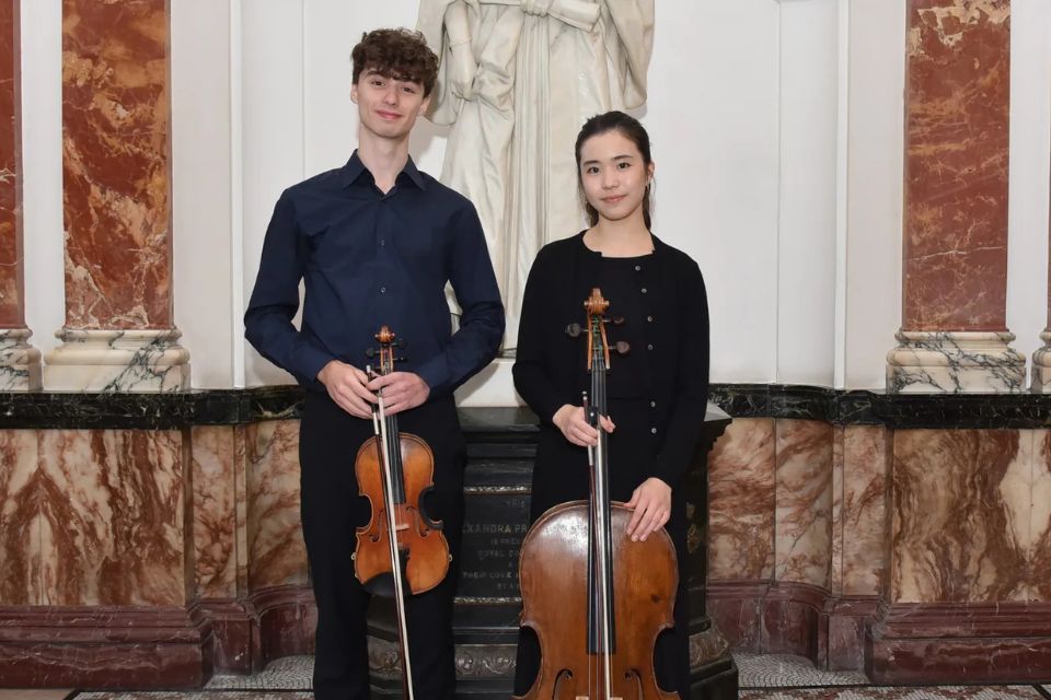 A boy and girl stand side by side wearing black and holding a violin and cello, respectively