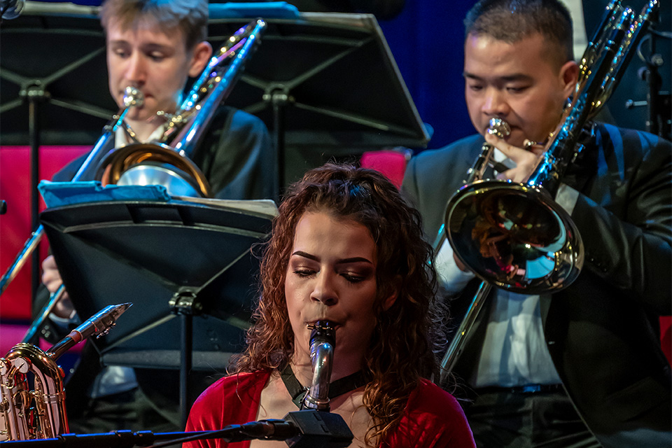 Three musicians play in the RCM Jazz Orchestra: a woman in a red top plays the saxophone in the foreground, and two men in suits play trombones in the background