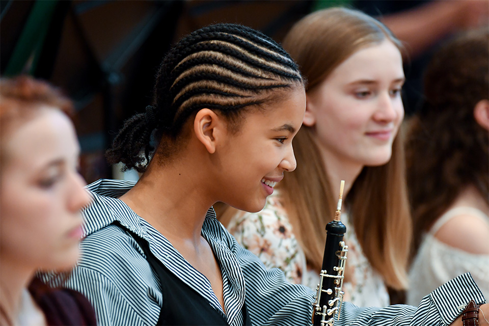 A girl from the Junior Department sits in an orchestra holding her oboe and smiling