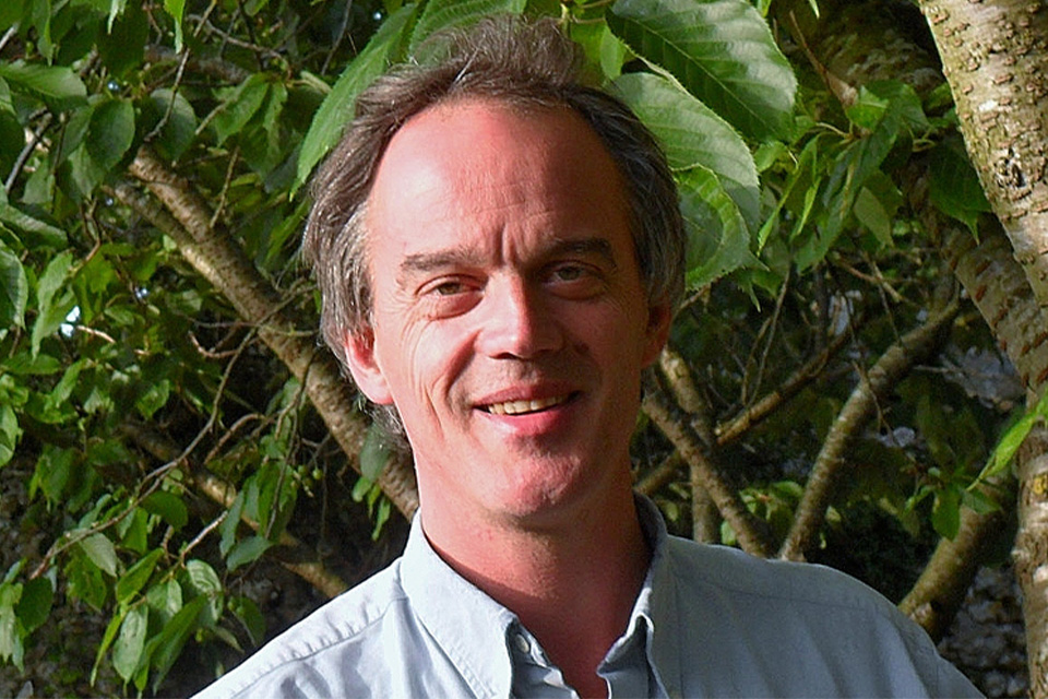 Richard Hosford stands outside against a green leafy backdrop, wearing a light blue shirt and smiling