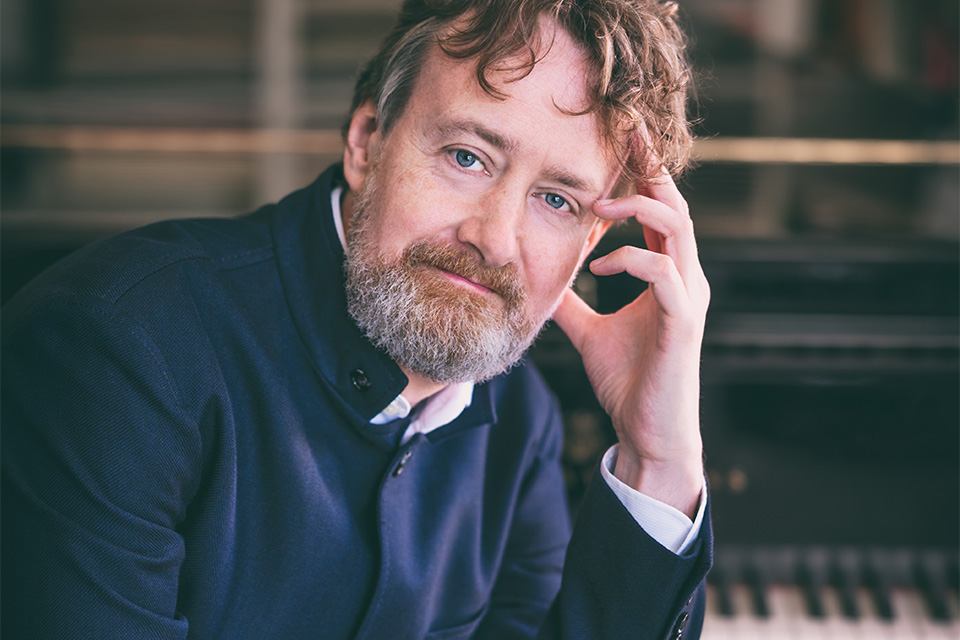 Roland Krüger wears a navy shirt and leans on a piano