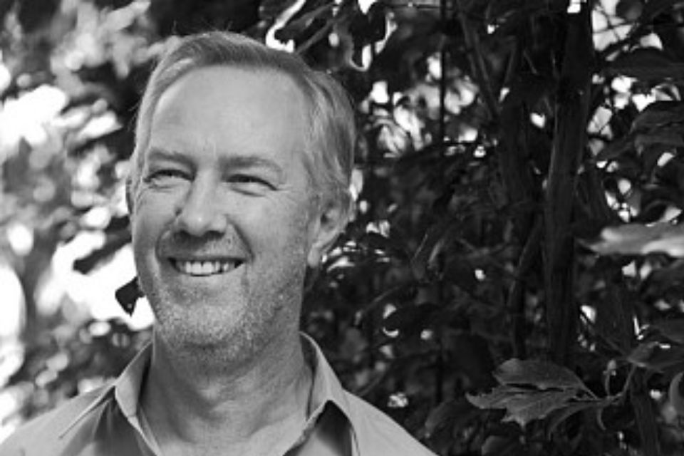 Roy Howat stands outside against a leafy background and looks to the side, smiling broadly