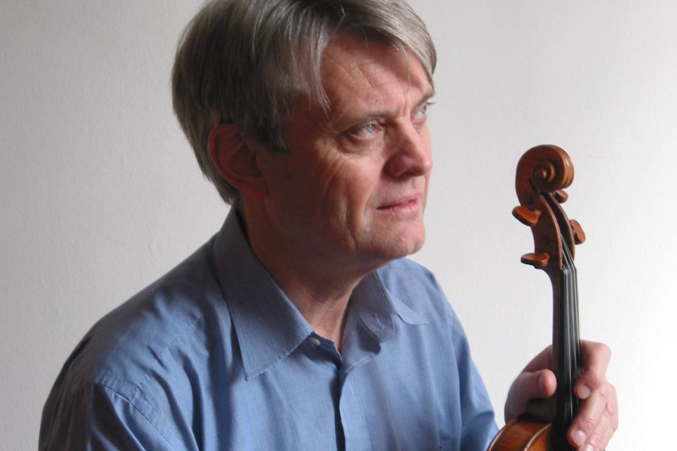 Simon Standage wears a blue shirt and holds his violin, looking up to the side