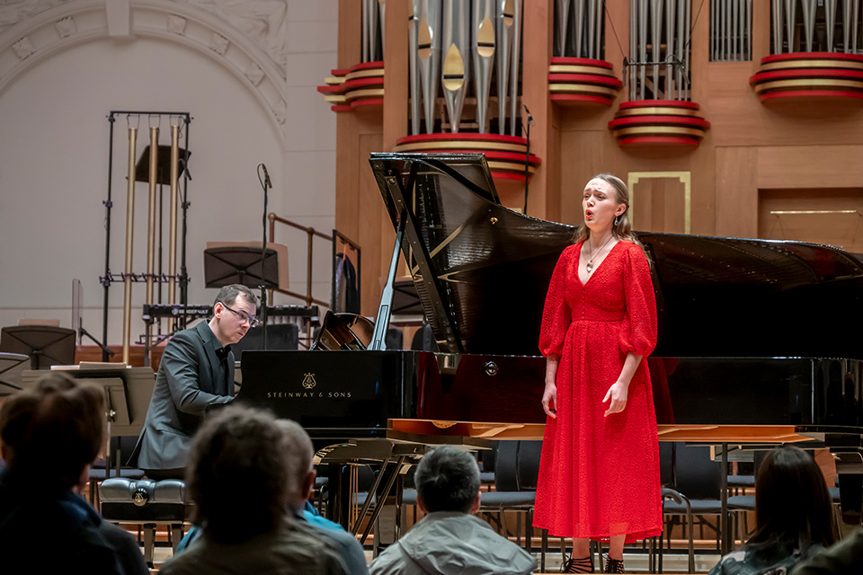 A woman in a red dress sings on stage to an audience, a piano accompanist behind her