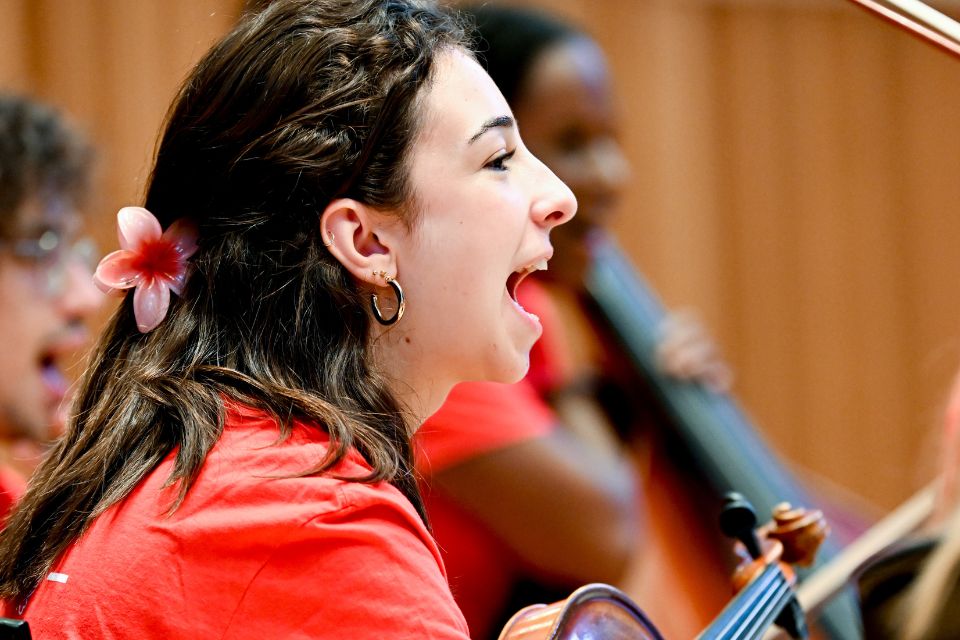 A girl wearing a red top holds her violin on stage next to other musicians, calling out happily