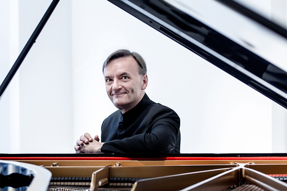 Stephen Hough, wearing black, sits at the piano stool and smiles at the camera through the piano lid