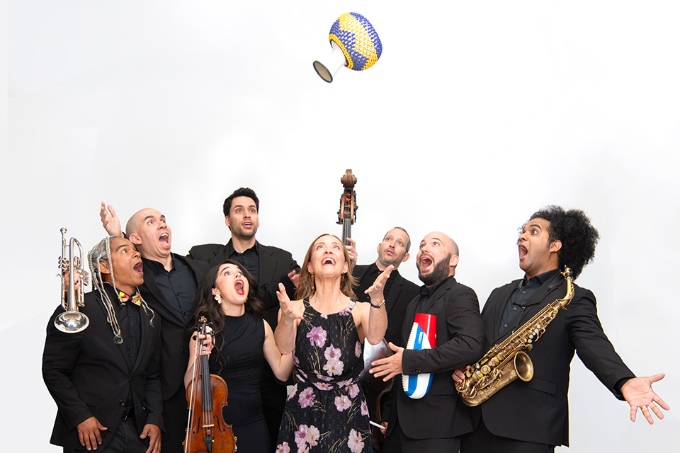 A group of people stand watching Sarah throw a shekere up in the air