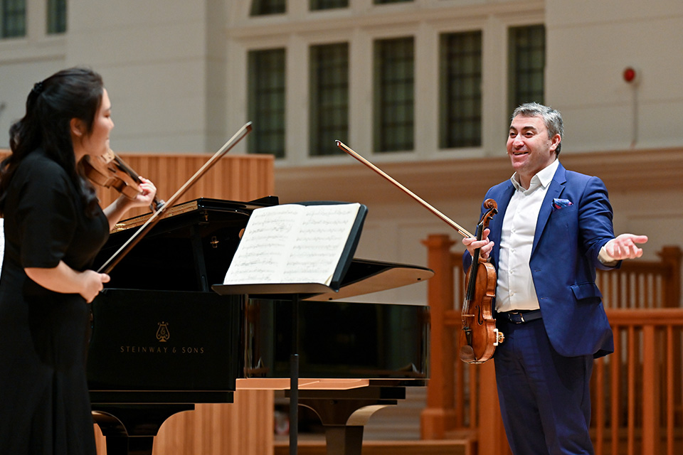 Maxim Vengerov holds his violin on stage, smiling at a violin student who he is teaching in a masterclass