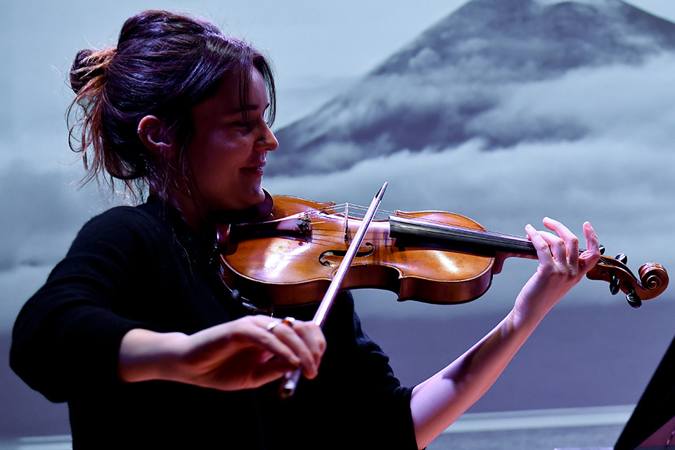 A young woman plays the violin with a projection of a mountain behind her