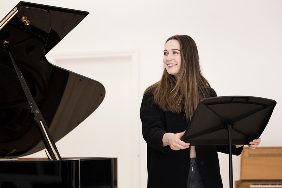A woman smiling, with brown hair, holding a music stand and looking to the side, with a piano behind her