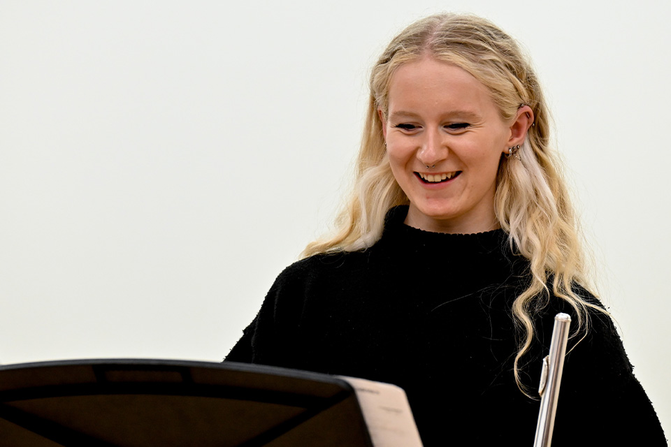 A young woman with blonde hair holds a flute and smiles