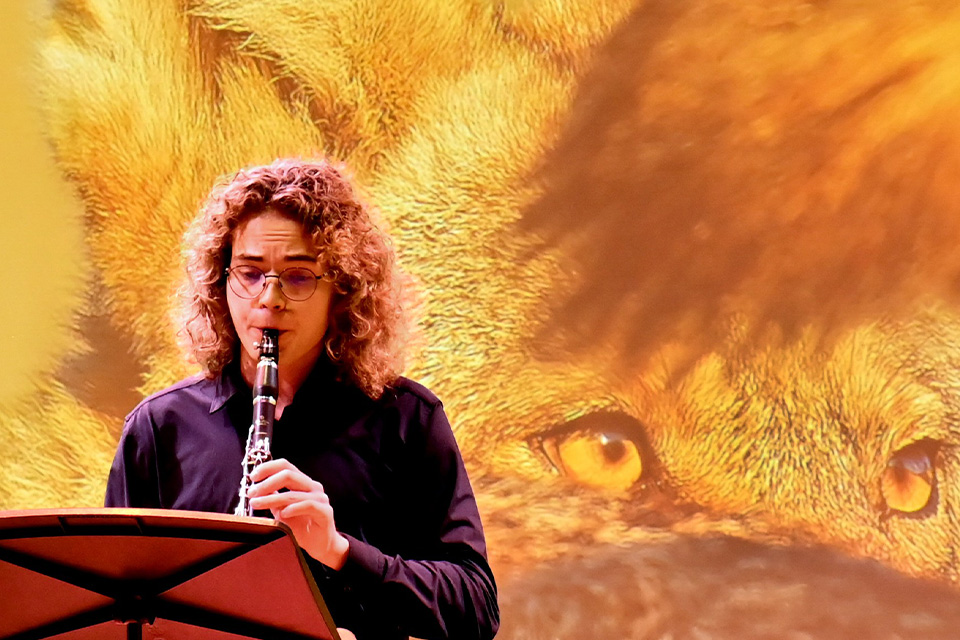 A young woman plays the clarinet in front of a projection of a fox's eyes