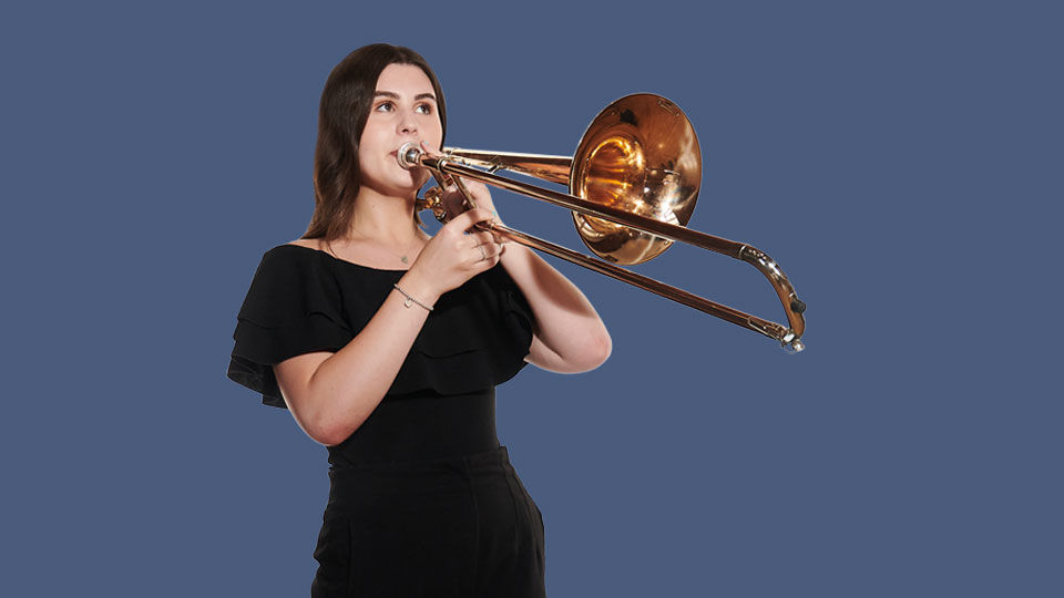 A female student, wearing a black dress, playing a trombone, with a indigo background.