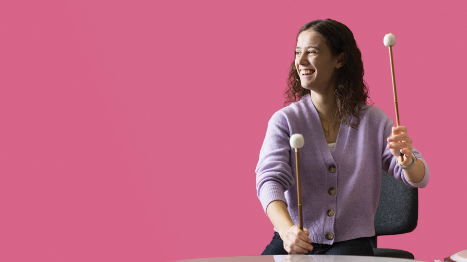 A women playing the timpani and looking to the side, with a deep, rose pink background.
