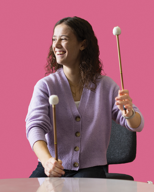 A women playing the timpani and looking to the side, with a deep, rose pink background.