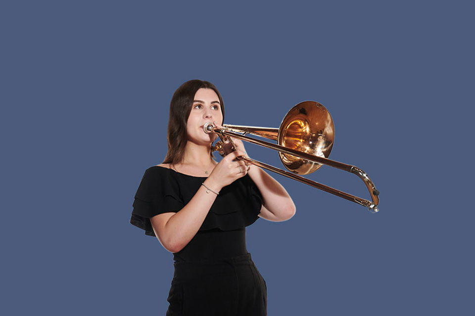 A female student, wearing a black dress, playing a trombone, with a indigo background.