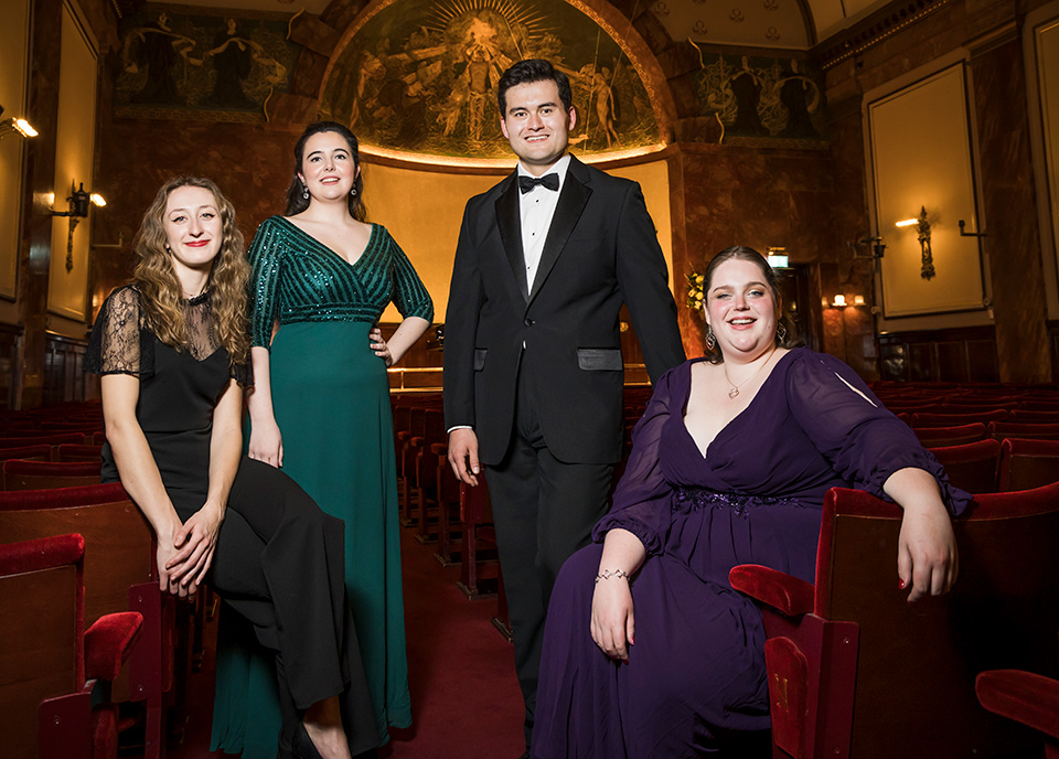 Three women and one man dressed in glamorous clothing in a grand room 