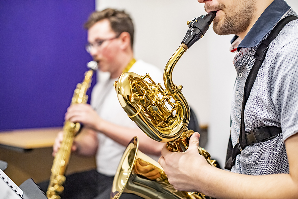 Two students, playing the tele saxophone and a baritone saxophone, in a well-lit practise room.