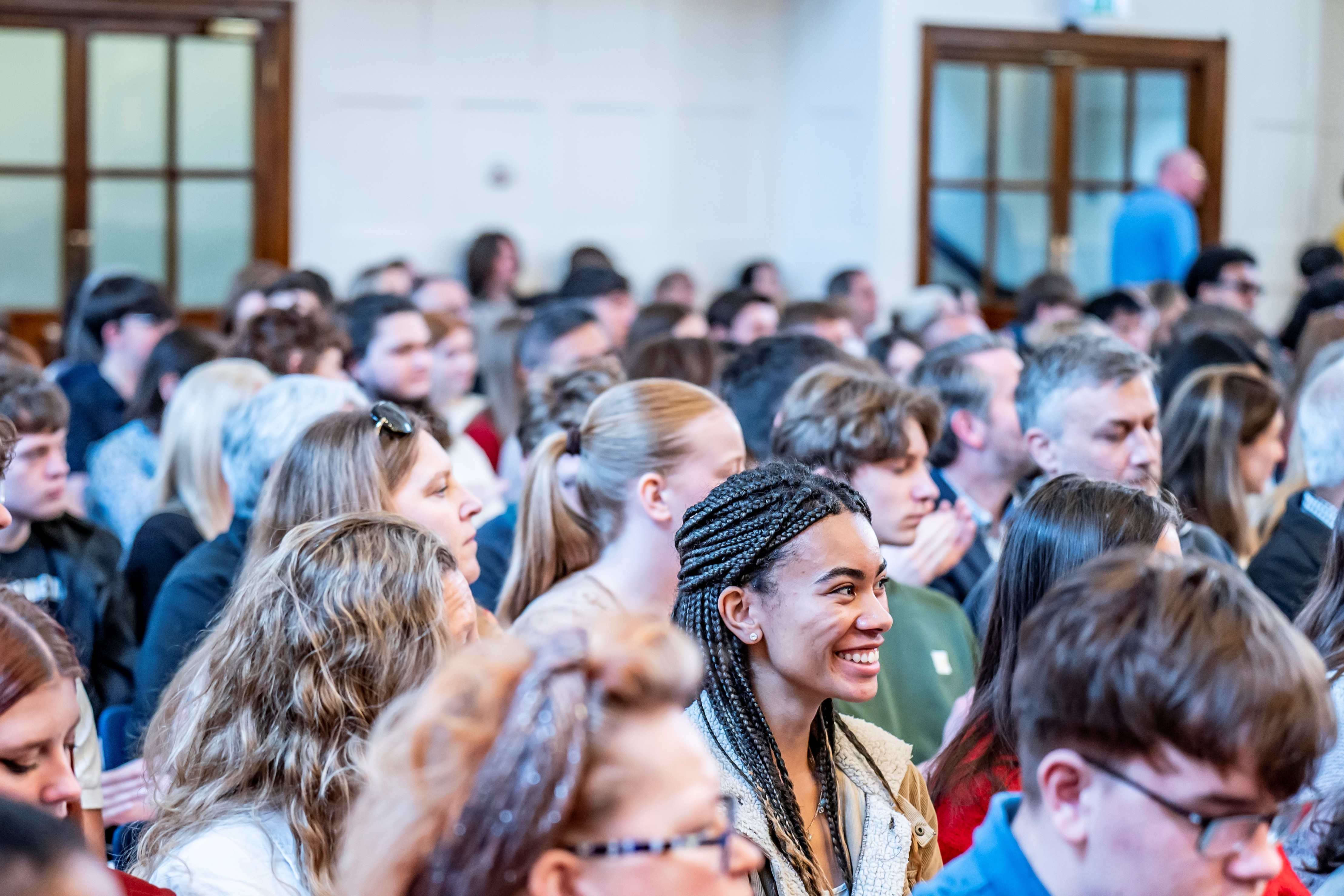 A large audience is seated closely together in a bright event hall, facing toward the front. In the foreground, a female individual with long, dark braided hair, wearing a light textured sweater and a jacket, appears to be smiling as they listen. They are surrounded by other attendees, some of whom are clapping. The room has tall windows and wooden-framed doors in the background.